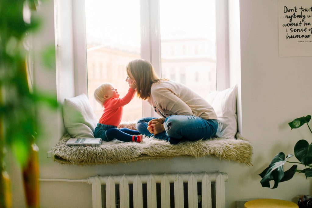 mother sitting in bay window area above a white furnace with a baby who is touching her face