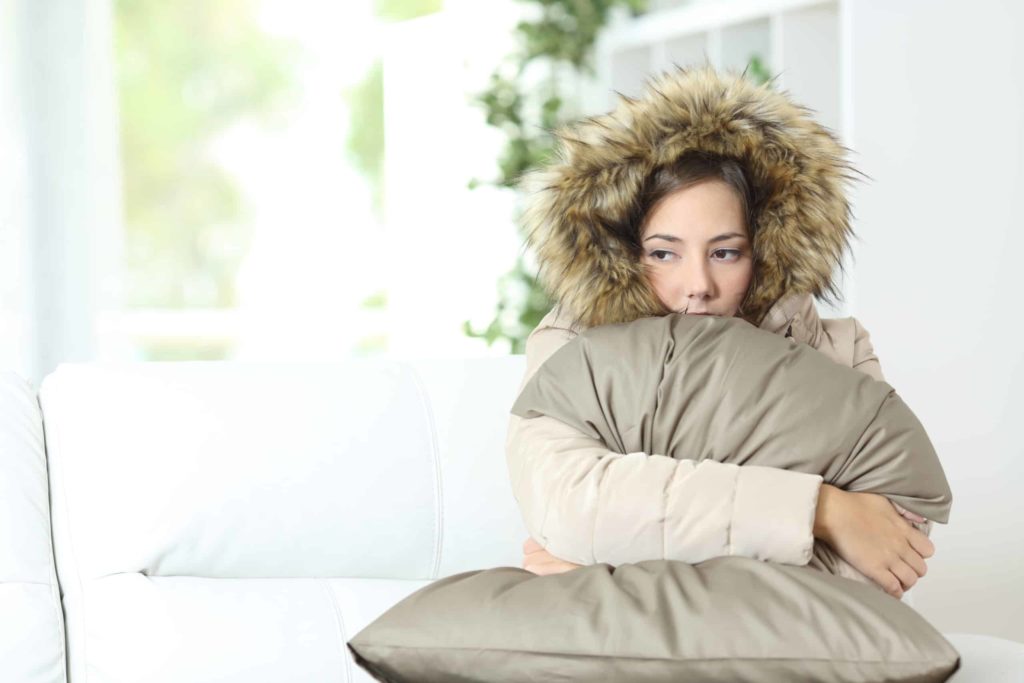 cold woman wearing a coat indoors while hugging a pillow to stay warm