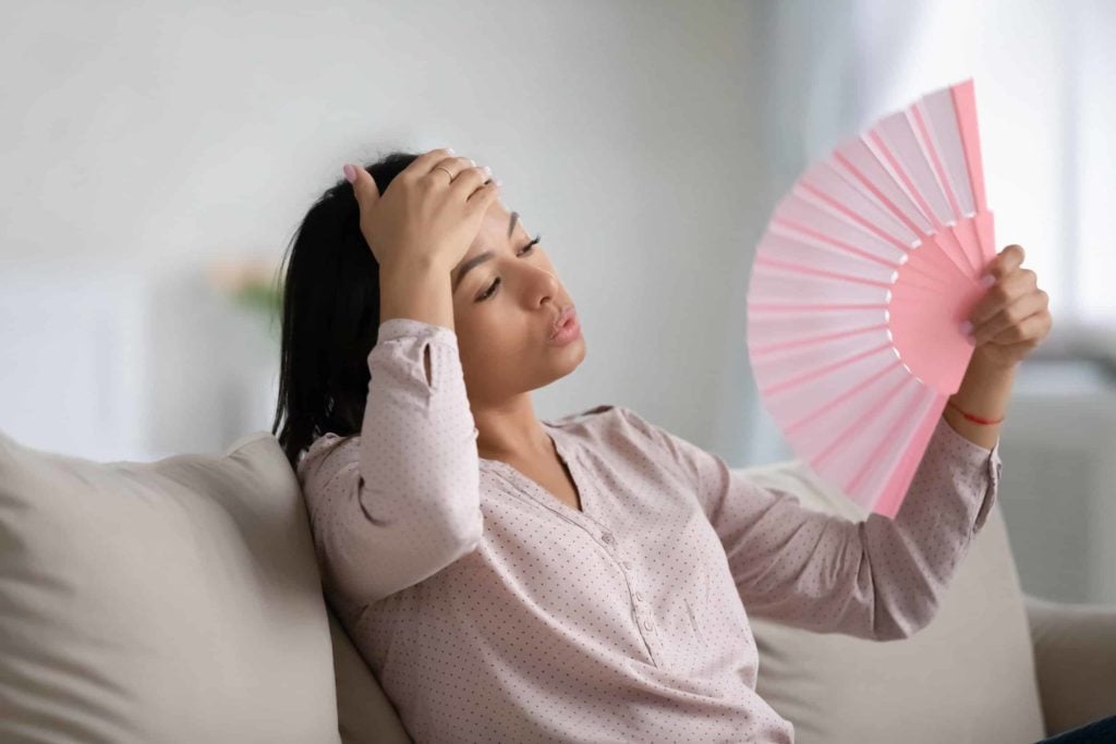 young woman fanning herself with pink fold out fan and her hand on her forehead hot because of broken ac